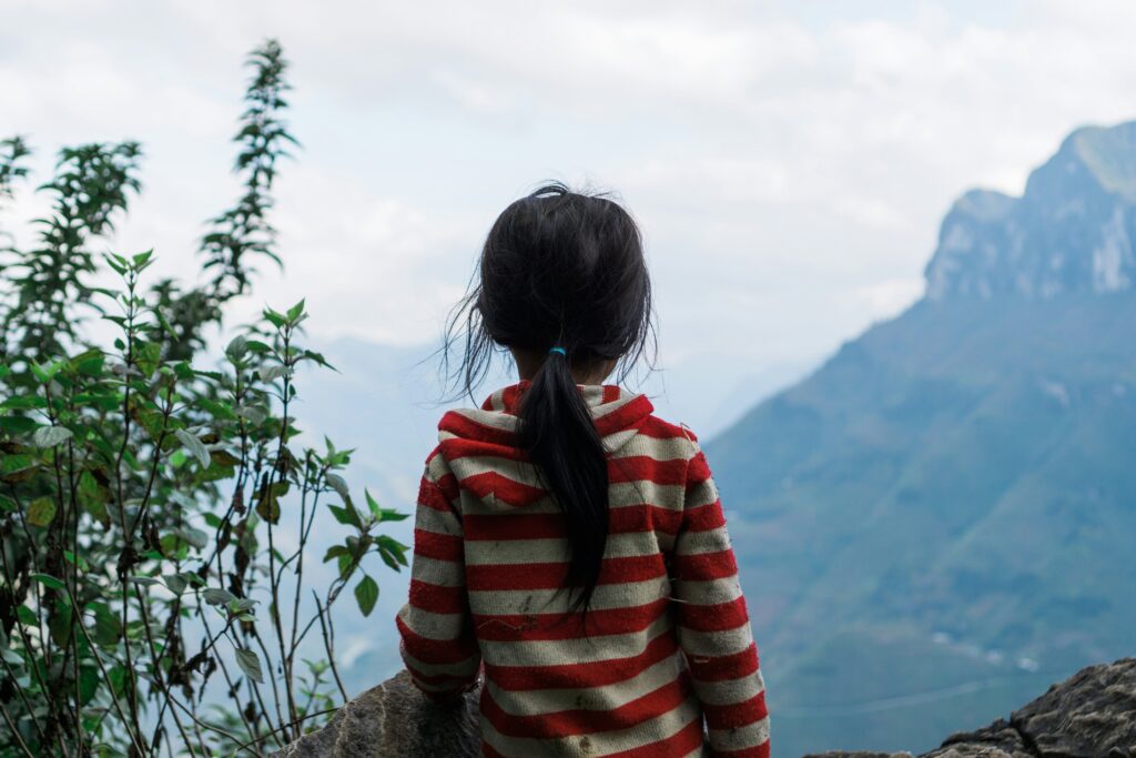 A girl with long hair wearing a red and cream striped sweater faces away looking out towards some mountain peaks.