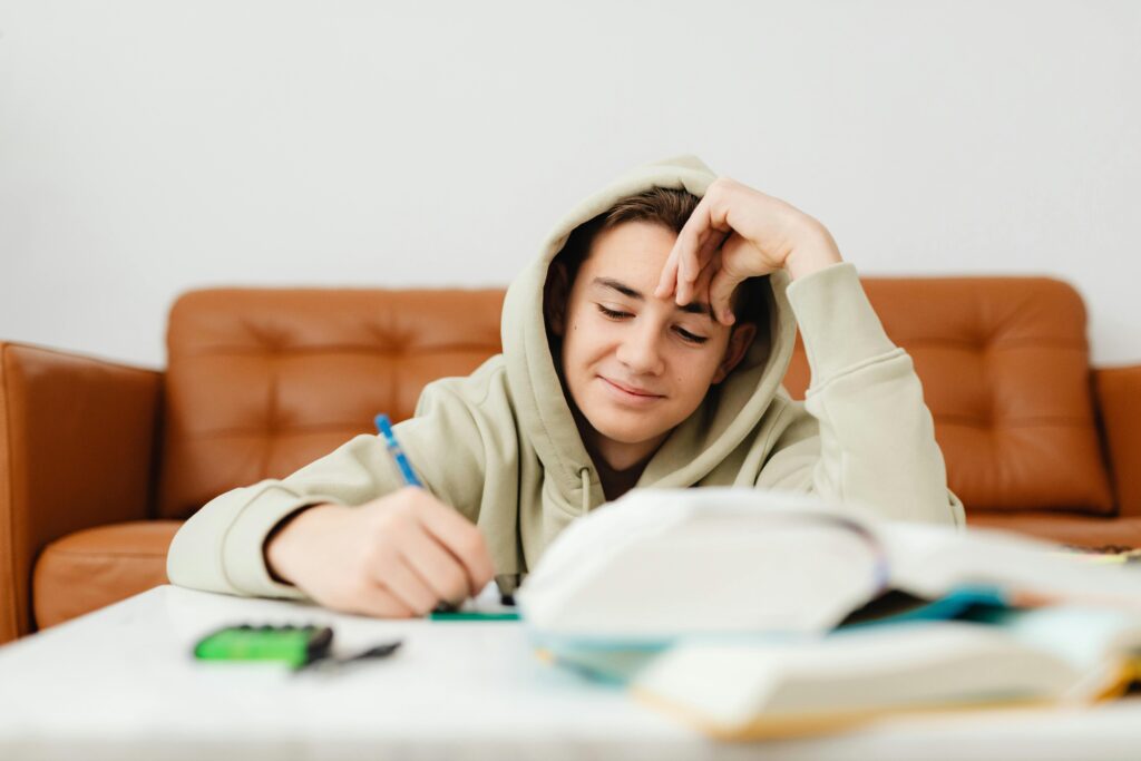 A boy wearing a sage green hooded sweatshirt has the hood over his brown hair and has one hand up to his forehead like he is thinking while his other hand holds a pencil. He writes on some paper on a table. Behind him is a brown leather couch.