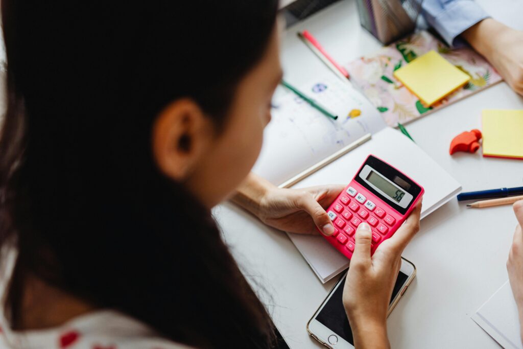 A young girl with long, dark hair holds a bright pink calculator. On the table in front of her lie notebooks, paper, yellow post-it notes, writing utensils and a pink eraser shaped like the top of a hammer.