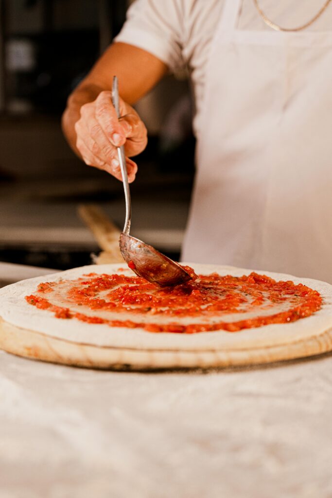A person wearing a white apron is using a metal ladle to spread tomato sauce in a spiral onto pizza dough that is rolled out on a wooden pizza launcher board.