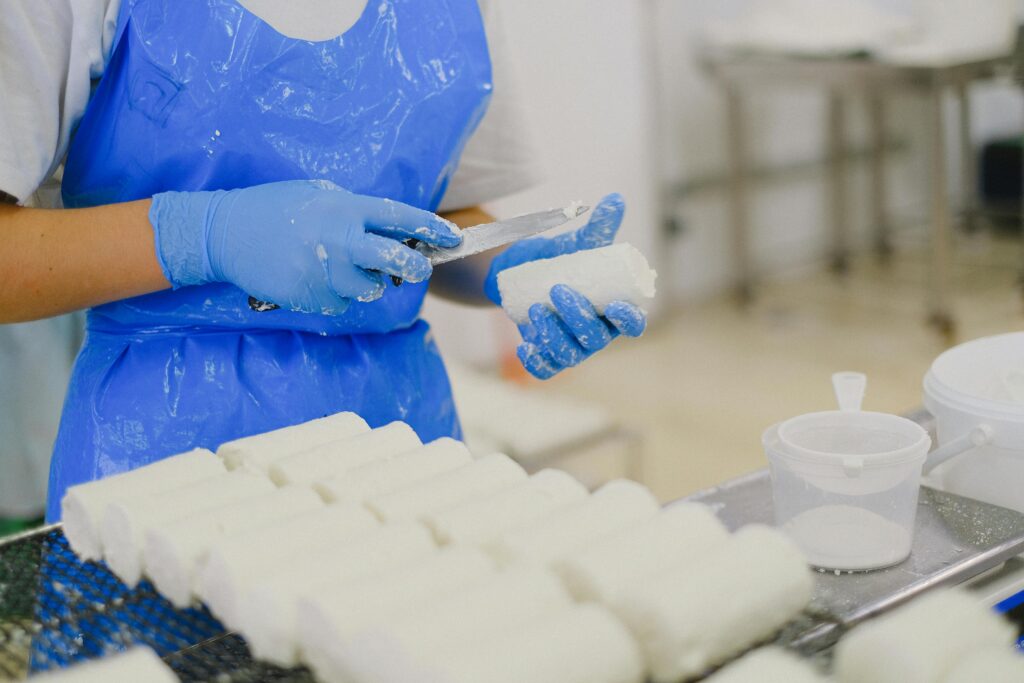 A person wears a blue apron and blue rubber gloves while holding a knife in one hand and a white log-shaped piece of food in the other hand. In front of them are two rows of the same food sitting on a metal rack.