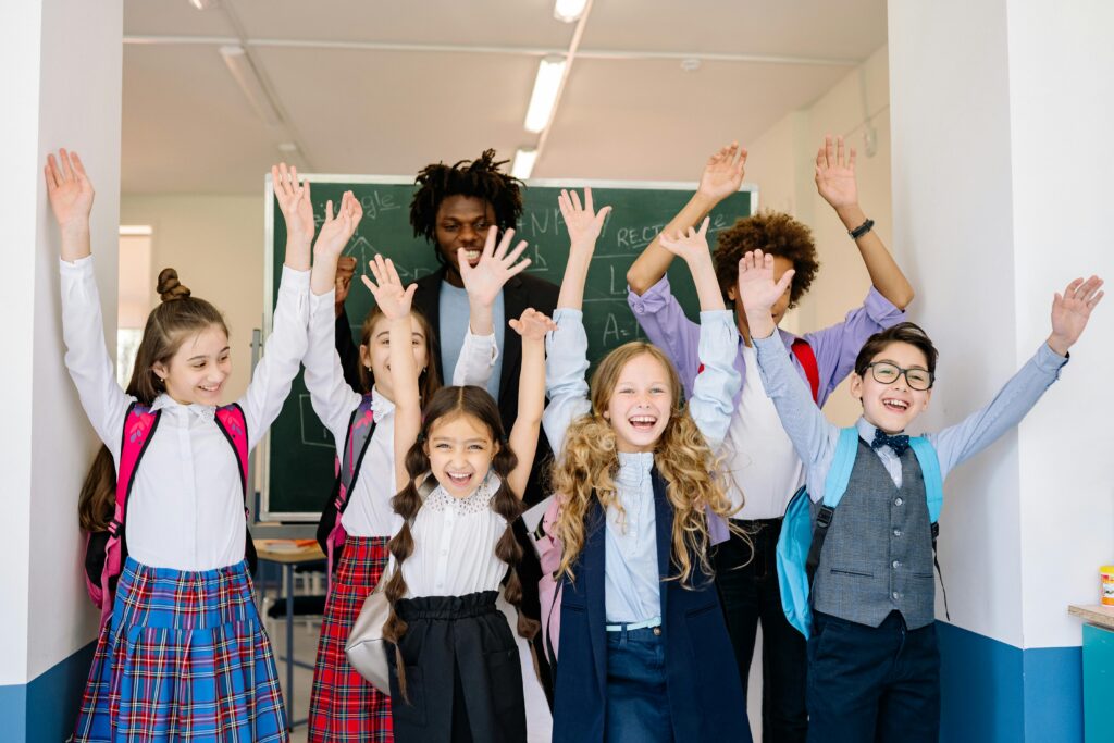 A group of six school children are smiling and holding their arms up in the air. The children are wearing dressy clothes and backpacks. Behind them is a green chalkboard and their teacher smiling.