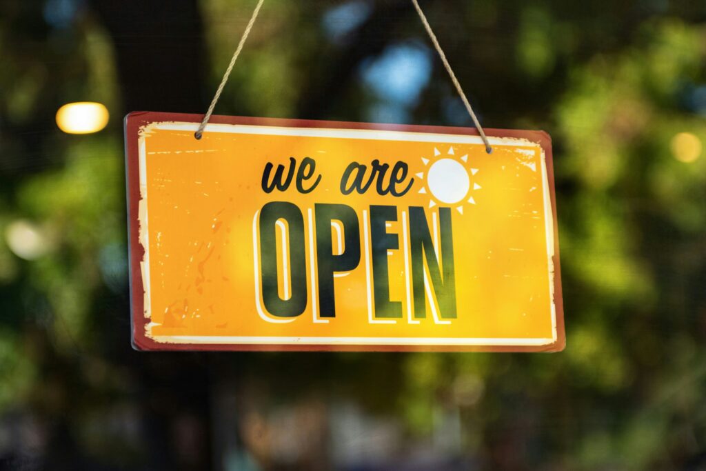 In the foreground is a wooden sign hanging by string. The sign has a yellow background with a white sun and words in green reading, "we are OPEN". In the backgrond are blurry green and brown leaves.