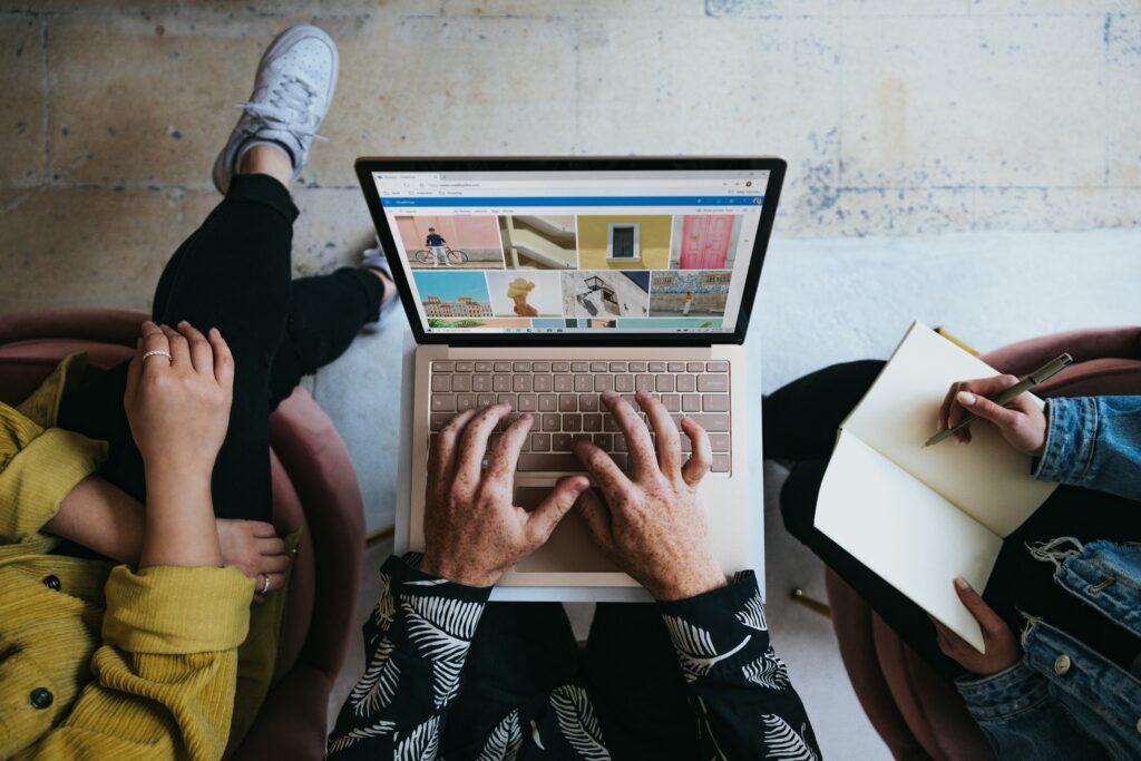 The photo is taken from above showing three people sitting around a laptop. The person in the middle has their hands on the keyboard and on the screen is a grid of colorful photographs. The person on the left is wearing a yellow jacket and has their legs crossed. The person on the right is holding a notebook and pen and appears to be taking notes.